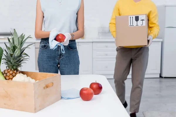 Cropped view of woman in latex gloves cleaning apple near boyfriend carrying carton box in kitchen — Photo de stock