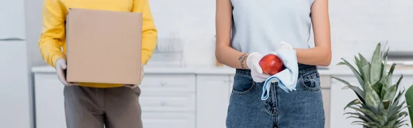 Cropped view of woman cleaning apple while boyfriend in latex gloves carrying carton box in kitchen, banner — Photo de stock