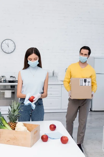 Man in medical mask holding carton box with signs near girlfriend cleaning apple and food in kitchen — стоковое фото