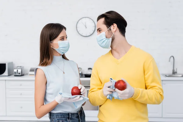 Woman in latex gloves cleaning apple near boyfriend in medical mask in kitchen — Stock Photo