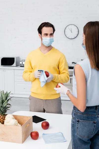 Man in medical mask cleaning apple with rag near girlfriend, food and digital tablet at home — Stock Photo