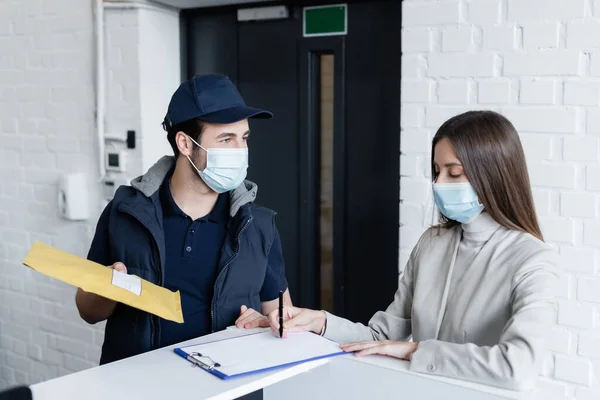 Businesswoman in medical mask writing on clipboard near courier with parcel in office — Stockfoto