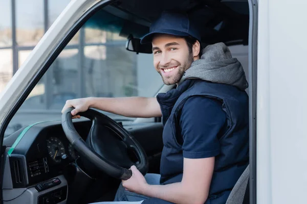 Smiling delivery man looking at camera near steering wheel in car — стоковое фото