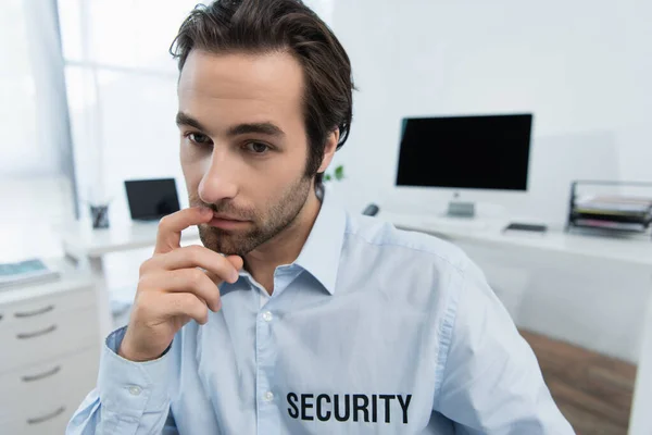 Nachdenklicher Wachmann im Hemd mit Sicherheitsaufdruck, der im Büro die Lippen berührt — Stockfoto