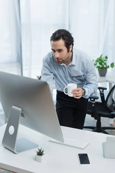 Jeune homme de sécurité avec tasse de thé regardant moniteur d'ordinateur dans la salle de sécurité — Photo de stock