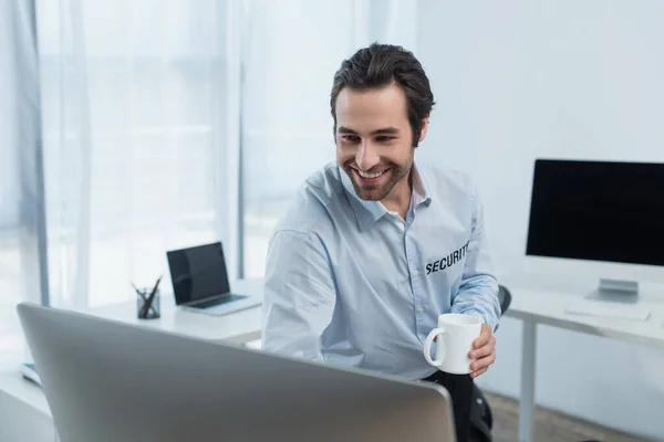 Jovem guarda com xícara de chá sorrindo perto de monitor de computador borrado na sala de segurança — Fotografia de Stock