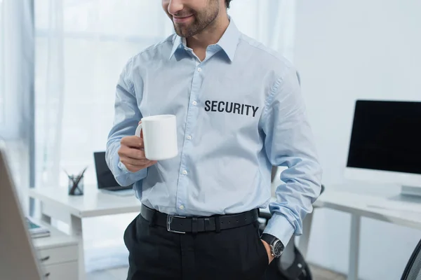 Обрізаний вид усміхненого чоловіка безпеки з чашкою чаю, що стоїть біля розмитих моніторів в офісі — стокове фото
