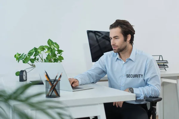 Hombre de seguridad en uniforme usando el ordenador portátil mientras trabaja en la sala de supervisión - foto de stock