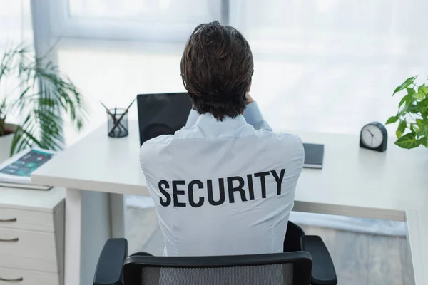 Vista trasera del guardia en camisa con letras de seguridad sentado cerca de la computadora portátil en la oficina - foto de stock