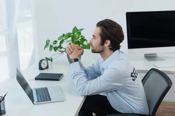 Side view of thoughtful security man sitting with clasped hands near laptop and monitor in supervision room — Stock Photo