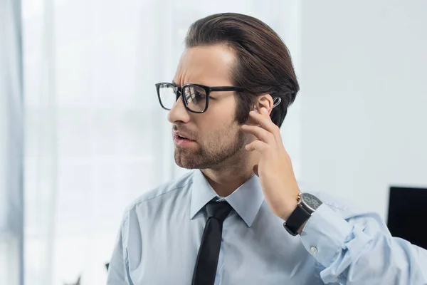 Seriöser Sicherheitsmann mit Brille, der bei der Arbeit im Büro Kopfhörer berührt — Stockfoto