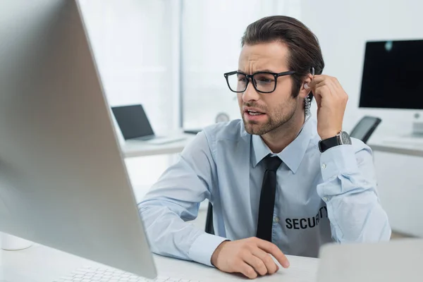 Guard in eyeglasses and earphone looking at computer monitor in security room — Stock Photo