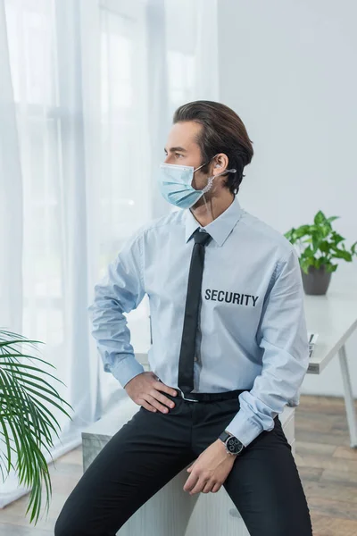 Security man in protective mask and earphone sitting on desk and looking away in office — Stock Photo