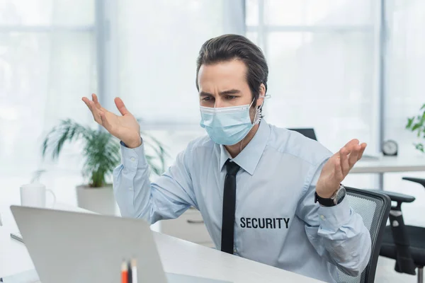 Homem de segurança desencorajado em máscara médica gestos perto de laptop turvo — Fotografia de Stock