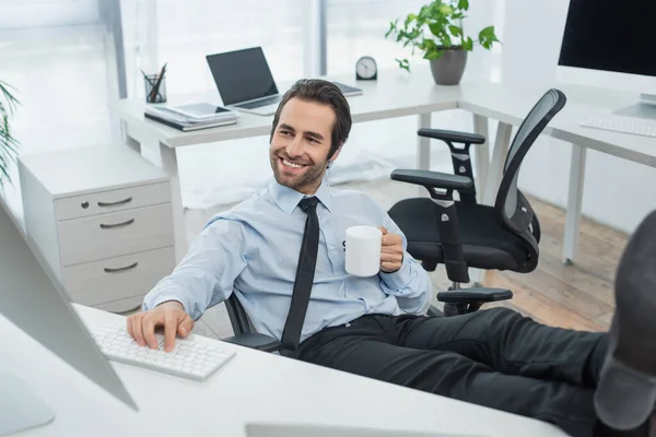 Fröhlicher Sicherheitsmann mit einer Tasse Tee, der auf den Monitor schaut, während er mit den Beinen auf dem Schreibtisch sitzt — Stockfoto