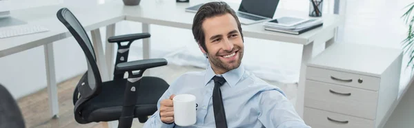 Alegre hombre de seguridad sosteniendo la taza de té mientras está sentado en la oficina, pancarta - foto de stock