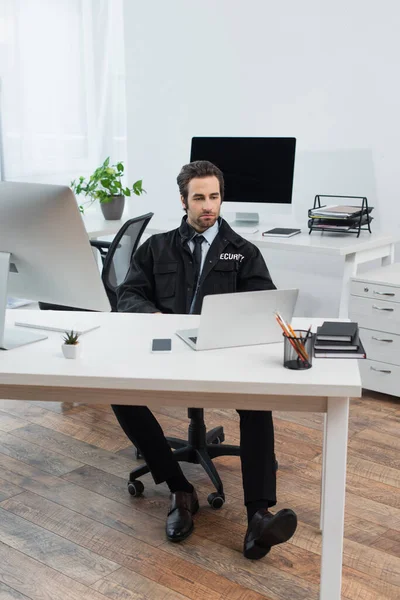 Sicherheitsmann schaut auf Laptop, während er am Arbeitsplatz im Überwachungsraum sitzt — Stockfoto