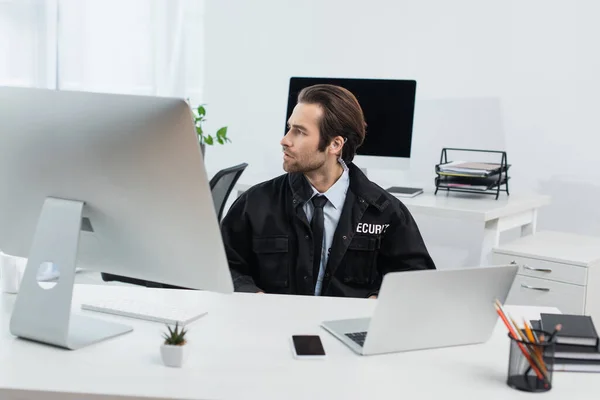 Hombre de seguridad sentado cerca de dispositivos en la sala de supervisión y mirando hacia otro lado - foto de stock