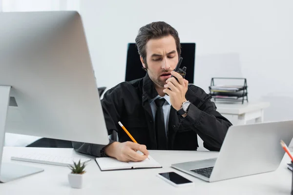 Hombre de seguridad hablando en walkie-talkie y escribiendo en un cuaderno cerca de computadoras en la oficina - foto de stock