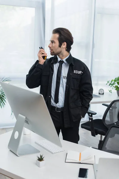 Guard in black uniform standing with hand in pocket at talking on walkie-talkie in office — Stock Photo