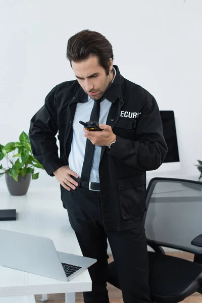 Homme de sécurité en uniforme noir appelant walkie-talkie tout en se tenant avec la main sur la hanche dans le bureau — Photo de stock