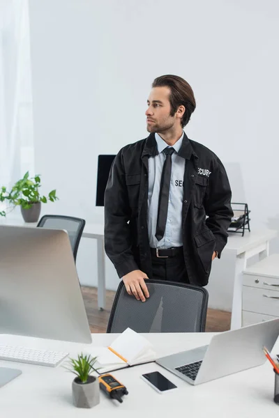 Seriöser Wachmann in schwarzer Uniform, der mit der Hand auf der Hüfte steht und in der Nähe von Computern wegschaut — Stockfoto