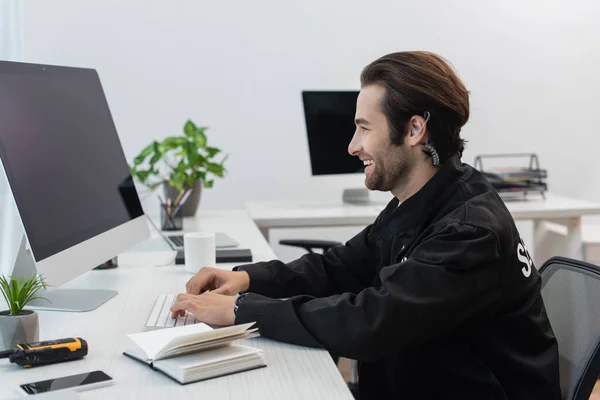 Vue latérale d'un homme de sécurité souriant tapant sur ordinateur près d'un ordinateur portable et talkie-walkie — Photo de stock