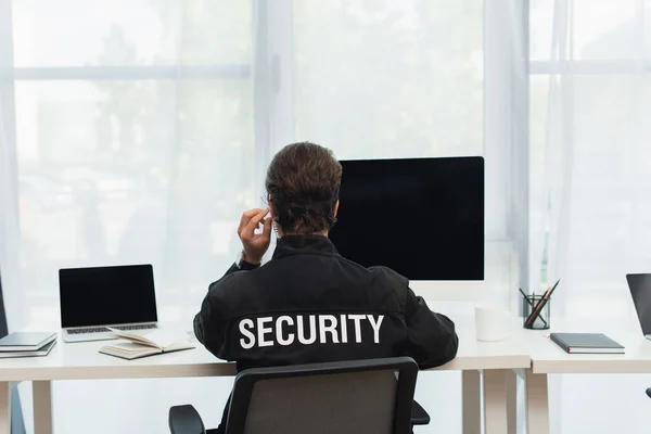 Vue arrière de l'homme de sécurité en uniforme noir et écouteur assis près des ordinateurs dans la salle de surveillance — Photo de stock