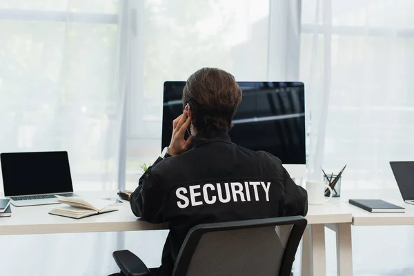 Rückansicht des Wachmannes in schwarzer Uniform mit Sicherheitsaufdruck, der neben Computern im Büro sitzt — Stockfoto