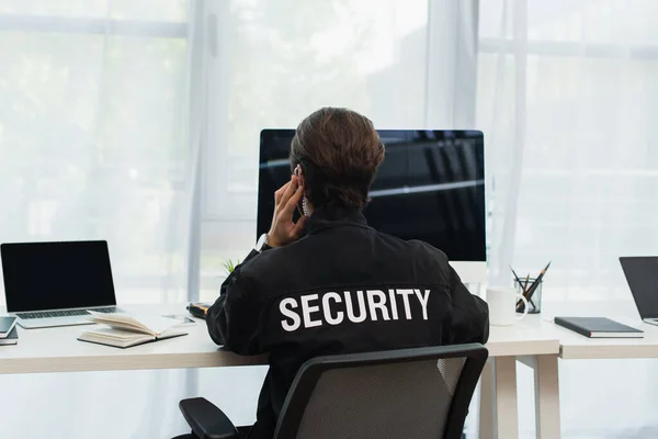 Vista trasera del guardia en uniforme negro con letras de seguridad sentado cerca de computadoras en la oficina - foto de stock