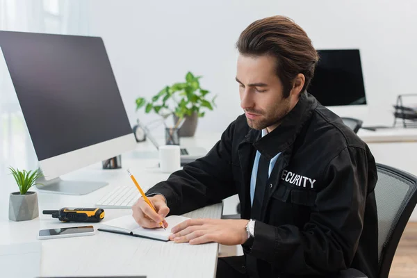 Hombre de seguridad escribiendo en el cuaderno cerca walkie-talkie y borrosa monitor de ordenador - foto de stock