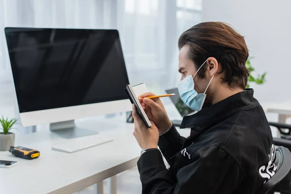 Sicherheitsmann in medizinischer Maske schreibt in Notizbuch neben verschwommenem Computermonitor — Stockfoto