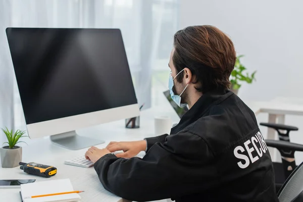 Sicherheitsmann in schwarzer Uniform und medizinischer Maske tippt im Überwachungsraum am Computer — Stockfoto