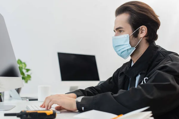 Seitenansicht des Wachmannes in medizinischer Maske, der am Computer in der Nähe von verschwommenem Walkie-Talkie arbeitet — Stockfoto