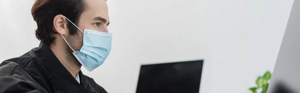 Vista laterale dell'uomo di sicurezza in maschera medica vicino a monitor di computer sfocati, banner — Foto stock