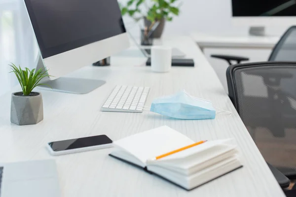 Notebook und medizinische Maske in Smartphone-Nähe mit leerem Bildschirm, Tastatur und Computermonitor — Stockfoto