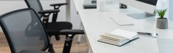 Notebook, mobile phone and flowerpot near keyboard on desk near blurred office chairs, banner — Stock Photo