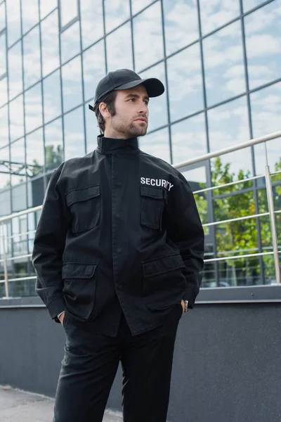 Jeune homme de sécurité debout avec les mains dans des poches d'uniforme noir et regardant loin à l'extérieur — Photo de stock