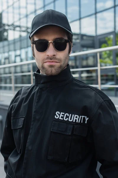 Охранник в черной форме и солнцезащитных очках смотрит в камеру на улице — стоковое фото