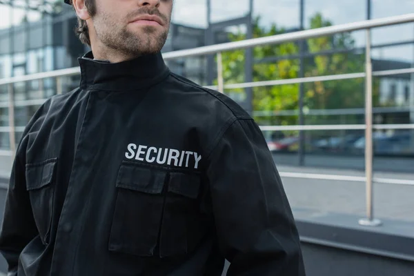 Visão parcial de guarda em uniforme preto com letras de segurança perto de edifício borrado — Fotografia de Stock