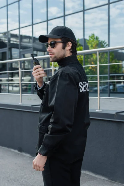 Young security man in uniform, cap and sunglasses talking on walkie-talkie near building — Stock Photo