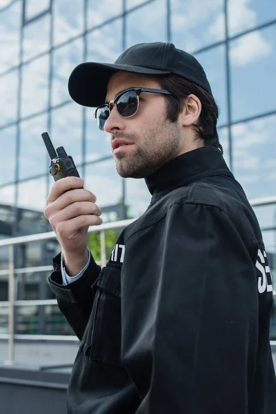 Serious guard in sunglasses and black uniform talking on radio set outdoors — Stock Photo