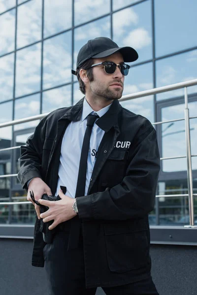 Giovane uomo di sicurezza in uniforme nera e occhiali da sole che prende la pistola dalla fondina sulla strada — Foto stock