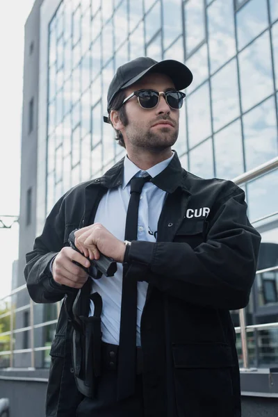 Guard in sunglasses and black uniform holding gun and looking away outdoors — Stock Photo