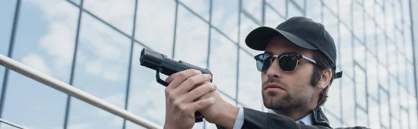 Joven hombre de seguridad en gorra negra y gafas de sol sosteniendo pistola al aire libre, pancarta — Stock Photo