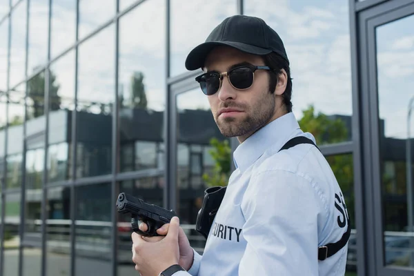 Security man in black cap and sunglasses holding gun outdoors — Stock Photo