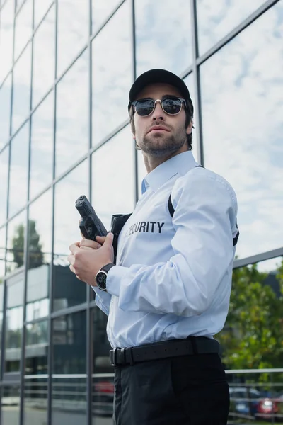 Young guard in sunglasses and black cap standing with gun near building with glass facade — Stock Photo