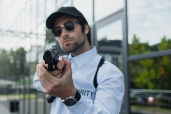 Blurred security man in cap and sunglasses holding gun outdoors — Stock Photo