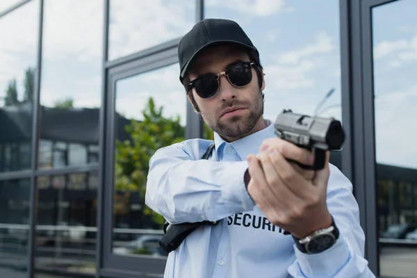 Joven guardia en uniforme, gorra negra y gafas de sol de pie con pistola al aire libre - foto de stock