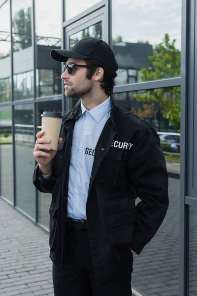 Giovane uomo di sicurezza in uniforme nera e cappuccio in piedi con caffè in strada — Foto stock